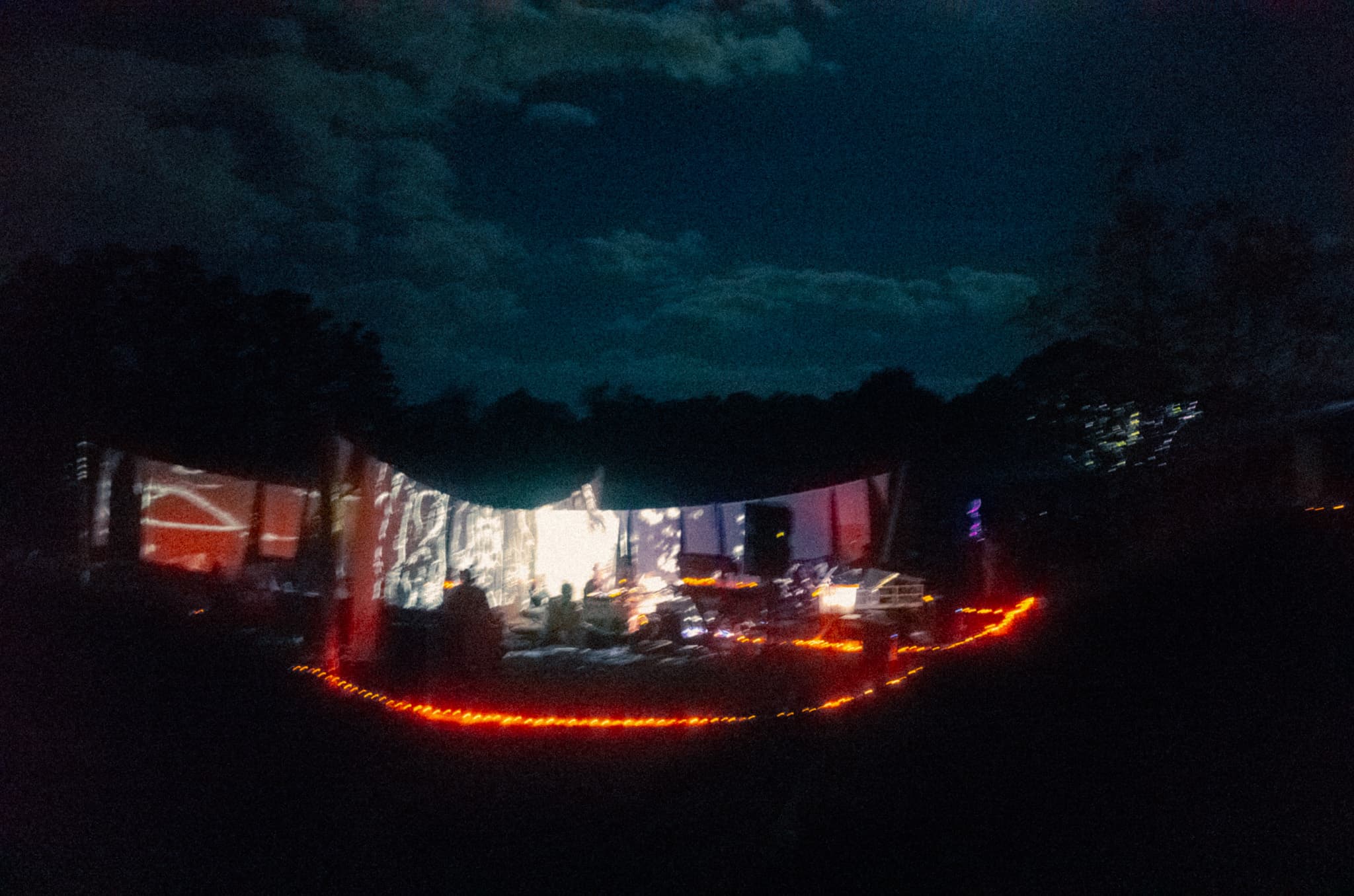 A DJ booth glowing under projected light in the night woods, string lights lining the path