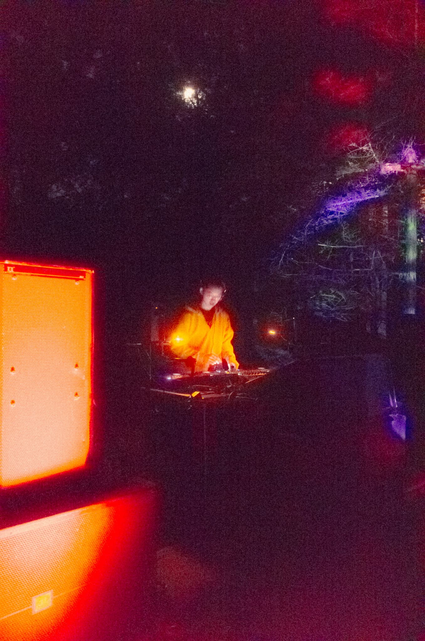 A DJ in a yellow jacket performing beneath the moon and colored lights in the trees