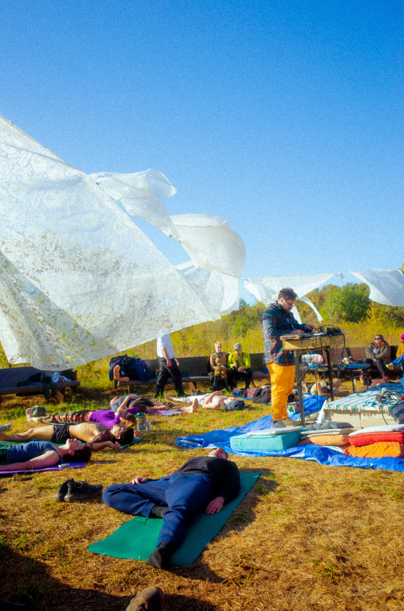 Sheets billowing over a group resting in the grass as a DJ performs at center
